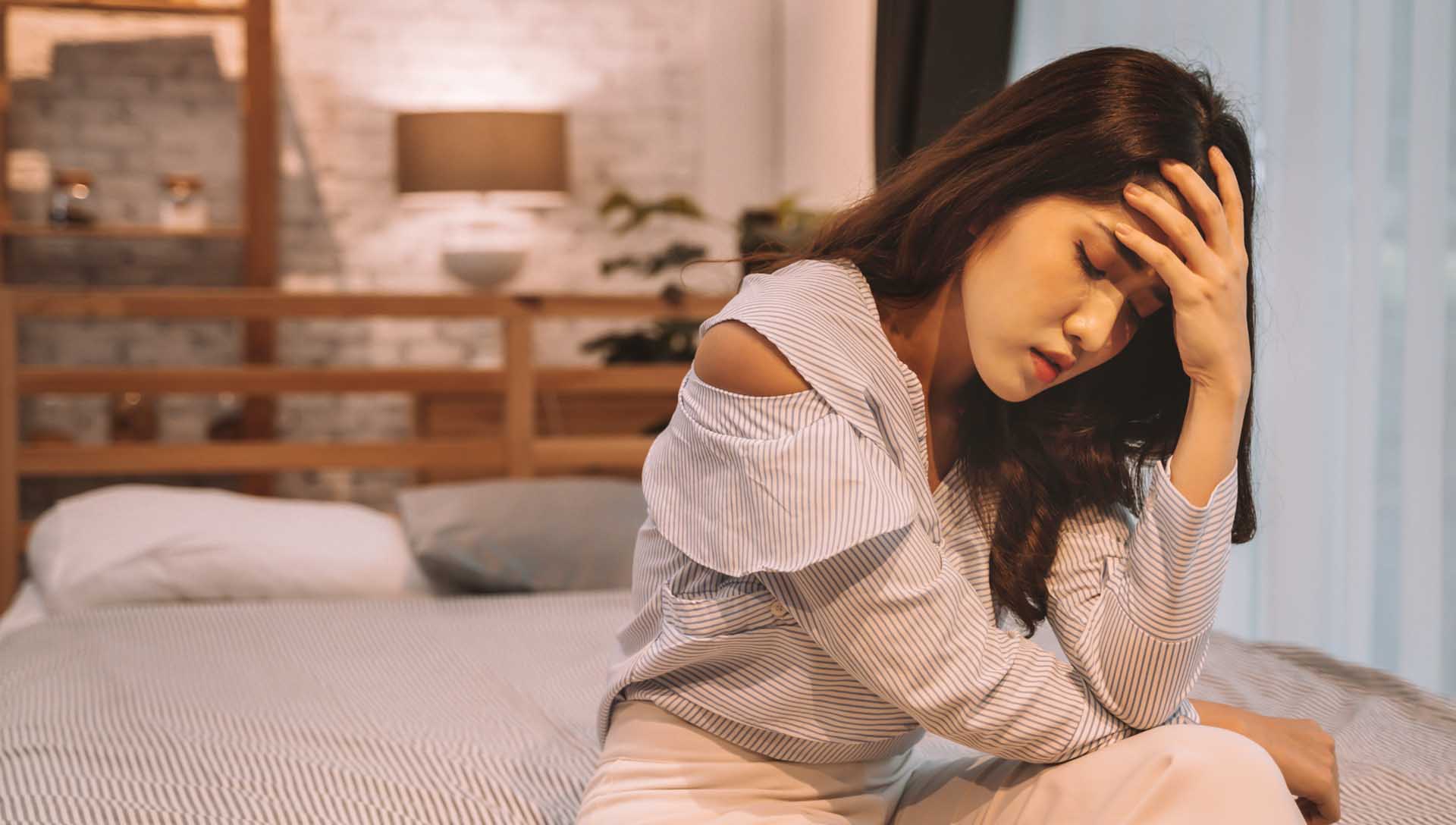 女性因頭痛與身體不適坐在床邊，手扶額頭呈現不舒服的狀態